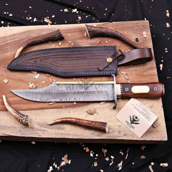 A custom handcrafted knife with a patterned steel blade, a brown wooden handle, and antler accents, displayed on a wooden board next to its leather sheath and a brand card.