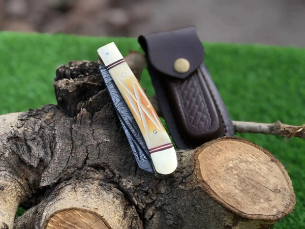 A folding pocket knife with a cream and orange inlay handle and blue-gray patterned blade, displayed next to a dark brown leather sheath on a rustic log with green artificial grass in the background.