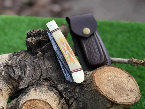 A folding pocket knife with a cream and orange inlay handle and blue-gray patterned blade, displayed next to a dark brown leather sheath on a rustic log with green artificial grass in the background.