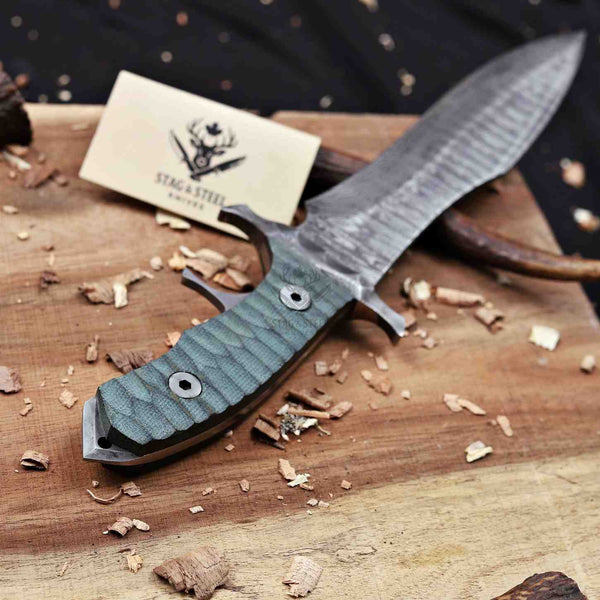A custom fixed-blade knife with a textured gray steel blade and a green-gray textured G10 handle, displayed on a wooden cutting board with wood shavings. A business card for ' Stag & Steel Knives' is visible in the background.