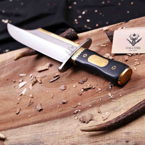 A fixed-blade knife with a satin-finished silver blade, brass guard and bolster, and a black handle with a brass accent, displayed on a rustic wooden board with wood shavings. A brand card for 'Stag & Steel Knives' is visible in the background.