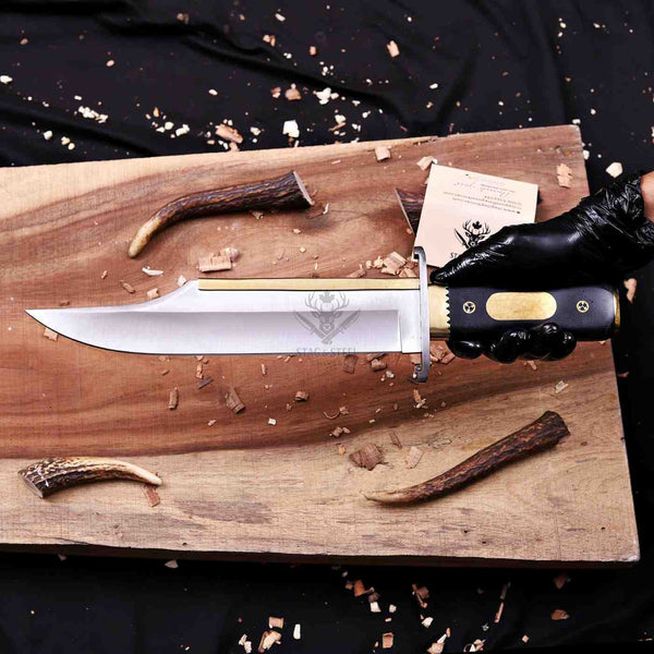A fixed-blade knife with a silver blade, gold bolster, and black handle is held by a black-gloved hand over a rustic wooden board with wood shavings and antler pieces.