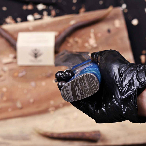 A hand wearing a black glove holds a carved wooden tool or handle with a blue and gray marbled inlay, set against a rustic wooden background with a blurred brand tag.