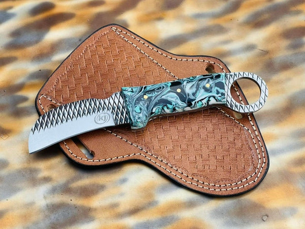 A fixed-blade knife with a textured silver blade and a marbled turquoise, green, and black handle, resting on a brown woven leather sheath over a leopard-print fabric background.
