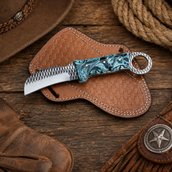 A fixed-blade Hawkbill knife with a resin handle, resting on a brown woven leather sheath over a rustic wooden surface with western-themed props.