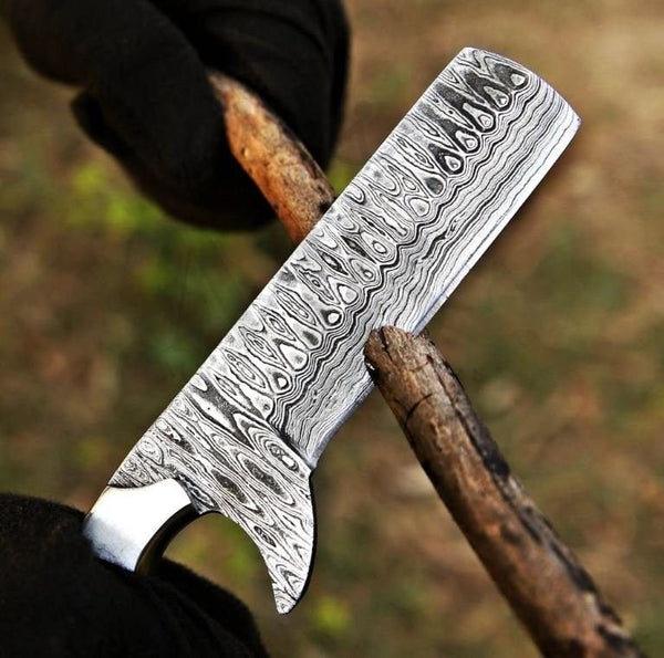 A close-up of a Damascus steel cleaver-style knife blade held upright, with a hand in a black glove gripping the wooden handle. The background is a blurred outdoor ground surface.