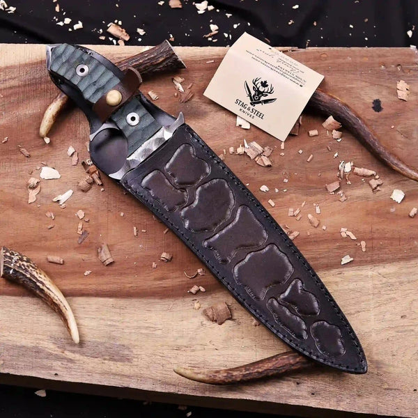 Embossed leather belt sheath holding tactical Damascus bowie knife staged on wooden workbench