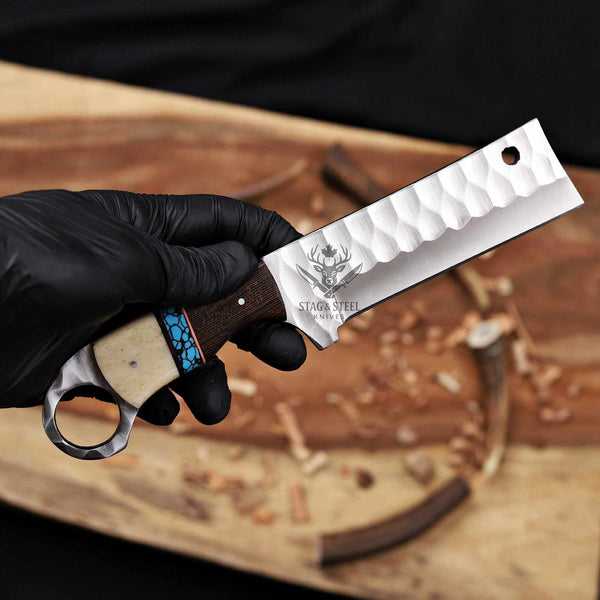 Hand holding a knife with a wooden handle and metal blade, branded 'Stag's Steel', against a workshop background.