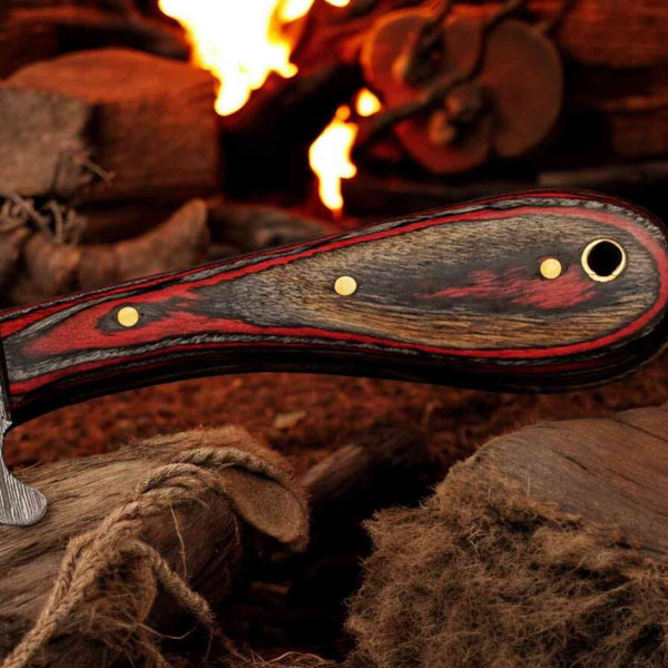 A close-up of a custom knife handle with a black and red wood grain pattern and brass pins, resting against rustic logs with a fire burning in the background.