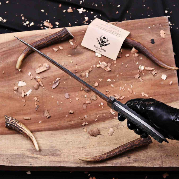 A hand wearing a black glove holds a long, dark knife over a rustic wooden cutting board scattered with wood shavings. Two antlers and a small branded card reading 'STOCK STEEL' are visible on the board.