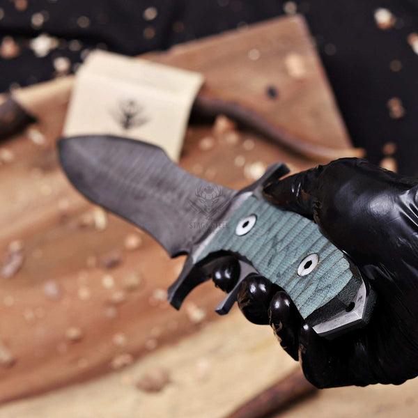 A close-up of a hand wearing a black glove holding a fixed-blade knife with a dark textured blade and a green textured handle, set against a rustic wooden background with wood shavings.