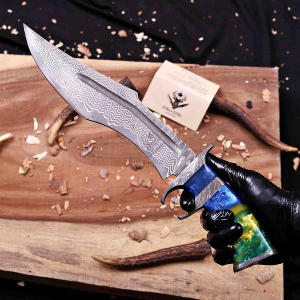 A hand wearing a black glove holds a fixed-blade knife with a patterned steel blade and a colorful handle. The knife is positioned over a wooden cutting board with wood shavings, antlers, and a small branded card in the background.
