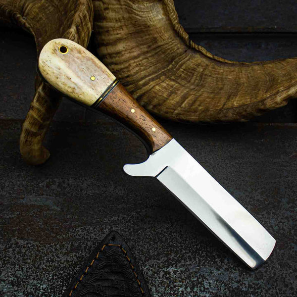 A fixed-blade knife with a polished silver blade, a dark brown wooden handle, and a natural beige horn or bone pommel, displayed diagonally on a dark textured surface with a horn prop in the background.