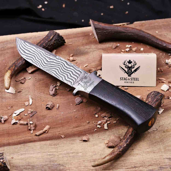 A fixed-blade knife with a patterned Damascus-style steel blade and a dark wood handle lies on a wooden surface with wood shavings and antlers around it. A small card reading Stag & Steel Knives is visible behind the knife.