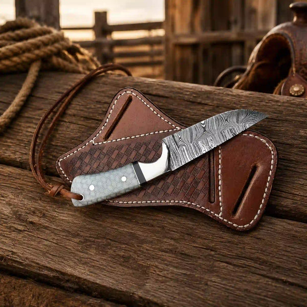 A fixed-blade knife with a patterned steel blade and textured handle rests on a brown leather sheath with a lanyard, placed on a rustic wooden surface with a rope and saddle in the background.