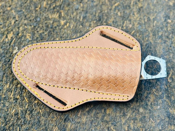 A tan leather knife sheath with a textured basketweave pattern, yellow stitching, and a silver metal keyring, shown on a dark textured background.