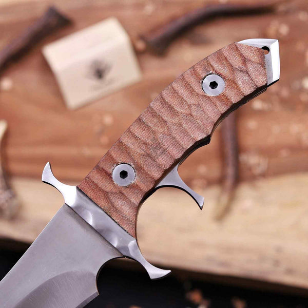 A close-up of a custom fixed-blade knife with a textured brown wooden handle and a silver steel blade, resting on a rustic wooden surface with blurred wood pieces in the background.