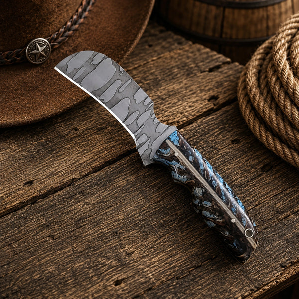 A custom knife with a patterned gray steel blade and a blue marbled handle, displayed diagonally on a rustic wooden surface next to a brown leather hat and a coil of rope.