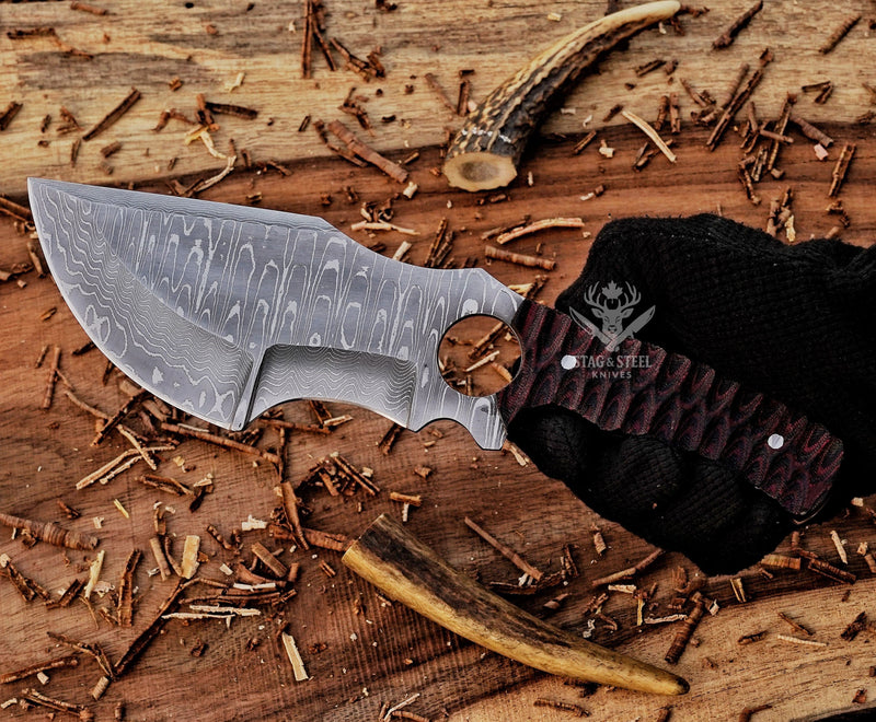 Handmade Damascus steel tracker knife with a textured dark handle, held in a gloved hand over wood shavings.