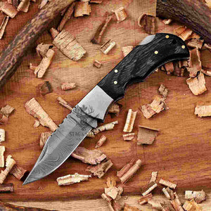 A Damascus Folding knife with a black Pakka wood handle, displayed diagonally on a wooden surface surrounded by wood shavings.