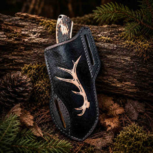 Black leather knife sheath featuring a laser-engraved tan antler design, resting on a rustic forest backdrop of pine needles and moss.