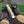 A folding pocket knife with a cream and orange inlay handle and blue-gray patterned blade, displayed next to a dark brown leather sheath on a rustic log with green artificial grass in the background.