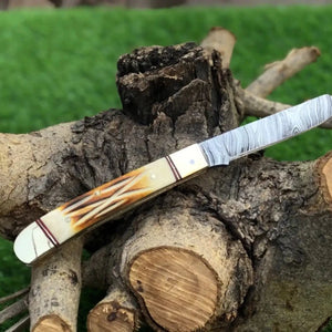 A custom pocket knife with a Damascus steel blade and a carved bone handle, displayed on a tree stump with a green grass background.