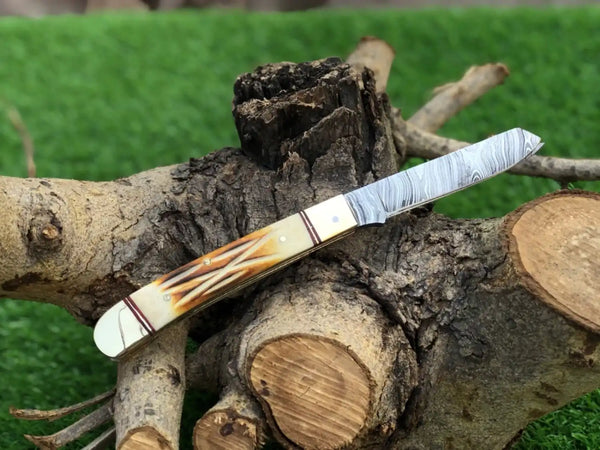 A custom pocket knife with a Damascus steel blade and a carved bone handle, displayed on a tree stump with a green grass background.