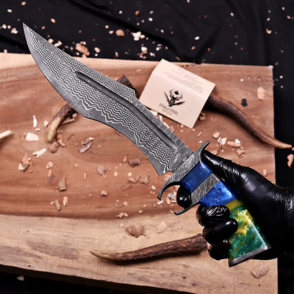 A hand wearing a black glove holds a San Mai Damascus Finger Guard Bowie knife over a wooden cutting board with wood shavings and a small branded card. The knife has a Stainless San Mai Damascus steel blade and a colorful handle with blue, green, and gold marbled sections.