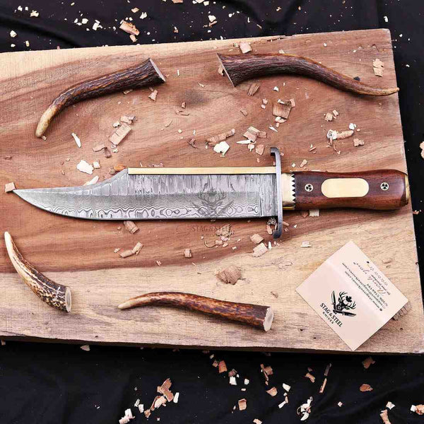 A custom handcrafted hunting knife with a Damascus steel blade and a dark wood handle, displayed on a wooden cutting board with antlers, wood shavings, and a brand card.