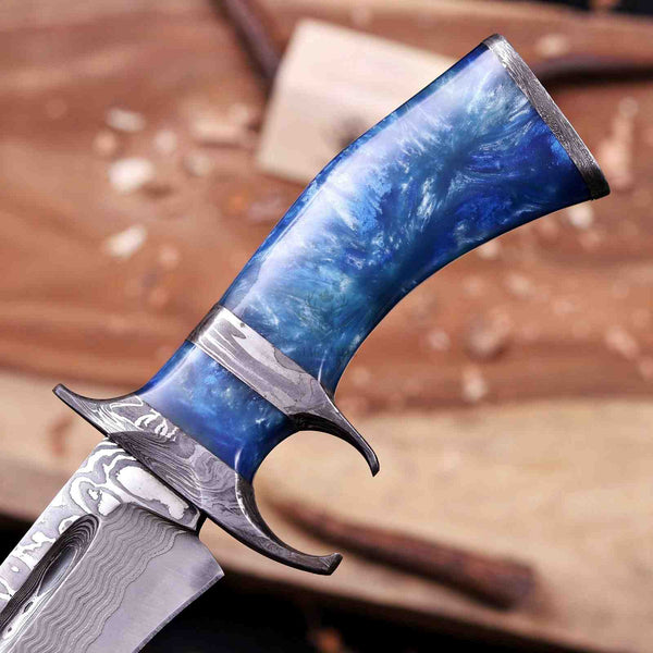 A close-up of a custom knife with a blue marbled resin handle and a Damascus steel blade, set against a blurred workshop background.