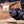 A hand wearing a black glove holds a carved wooden tool or handle with a blue and gray marbled inlay, set against a rustic wooden background with a blurred brand tag.