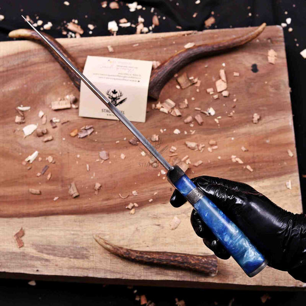 A hand wearing a black glove holds a long knife with a blue marbled handle over a wooden cutting board scattered with wood shavings. A small card and antler-like props are visible on the board.