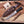 A fixed-blade knife with a patterned steel blade and black handle, shown with a brown leather sheath on a wooden surface. A small branded card reading 'The Knifestool Company' is visible, along with wood shavings and rustic wood props.