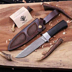 A fixed-blade knife with a patterned steel blade and black handle, shown with a brown leather sheath on a wooden surface. A small branded card reading 'The Knifestool Company' is visible, along with wood shavings and rustic wood props.