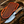 A fixed-blade knife with a patterned silver blade and a carved handle, shown next to its orange-brown leather sheath on a rustic wood surface with antlers and a pinecone.