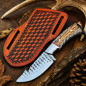 A fixed-blade knife with a patterned silver blade and a carved handle, shown next to its orange-brown leather sheath on a rustic wood surface with antlers and a pinecone.