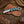 A fixed-blade Hawkbill knife with a resin handle, resting on a brown woven leather sheath over a rustic wooden surface with western-themed props.