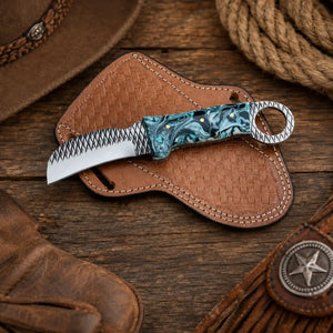 A fixed-blade Hawkbill knife with a resin handle, resting on a brown woven leather sheath over a rustic wooden surface with western-themed props.