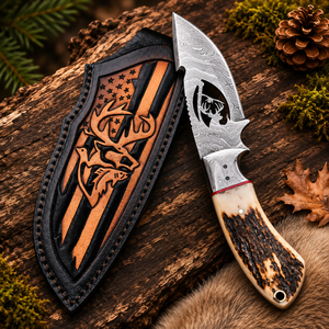 Stag horn Damascus gut hook hunting knife with skull cutout blade, wire notch, and matching laser-engraved stag leather sheath on a rustic forest log backdrop.