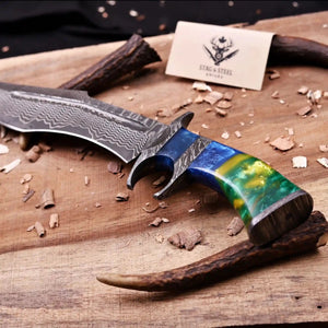 A custom San Mai Damascus steel Bowie knife with a vibrant blue, green, and yellow marbled handle, displayed on a rustic wooden board with wood shavings and antler props. A brand card for 'Stag Steel Knives' is visible in the background.