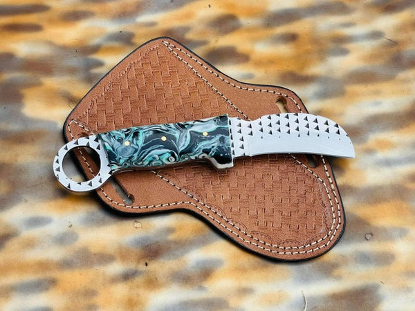 A custom folding pocket knife with a marbled green and blue handle, a silver studded blade, and a silver ring pommel, resting on its brown woven leather sheath over a leopard print background.