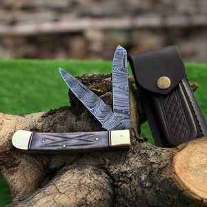 Two pocket knives with Damascus steel blades and purple handles on a wooden log, with a leather sheath in the background.