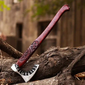 A hand-crafted Viking-style axe with a carved reddish-brown wooden handle and a Carbon Steel head featuring black runic engravings, resting on a log outdoors.