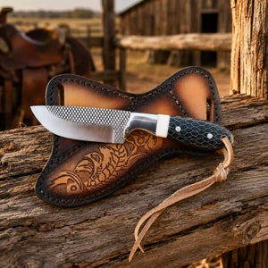A custom knife with a textured silver blade and a black honeycomb-patterned handle, resting on a brown leather sheath with floral tooling, set on a rustic wooden fence in a western-themed outdoor environment.