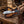 A knife with a patterned silver blade and dark handle resting in a brown tooled leather sheath on a rustic wooden surface, with a leather saddle and rope nearby.
