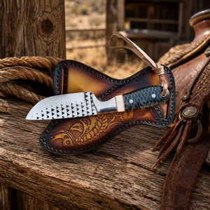 A knife with a patterned silver blade and dark handle resting in a brown tooled leather sheath on a rustic wooden surface, with a leather saddle and rope nearby.