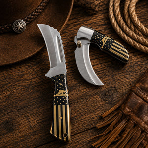 Two curved pocket knives with black and gold American flag patterned handles are displayed on a rustic wooden surface alongside a brown leather hat, rope, and fringed leather accessory.