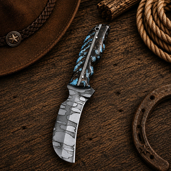 A custom knife with a patterned gray and silver blade and a blue/black marbled handle, displayed on a rustic wooden surface with a cowboy hat, rope, and horseshoe as props.
