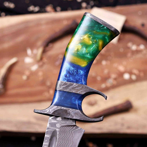 A close-up of a custom knife with a patterned Damascus steel blade and a highly polished handle featuring a gradient of blue, green, and yellow, resting on a rustic wooden surface.